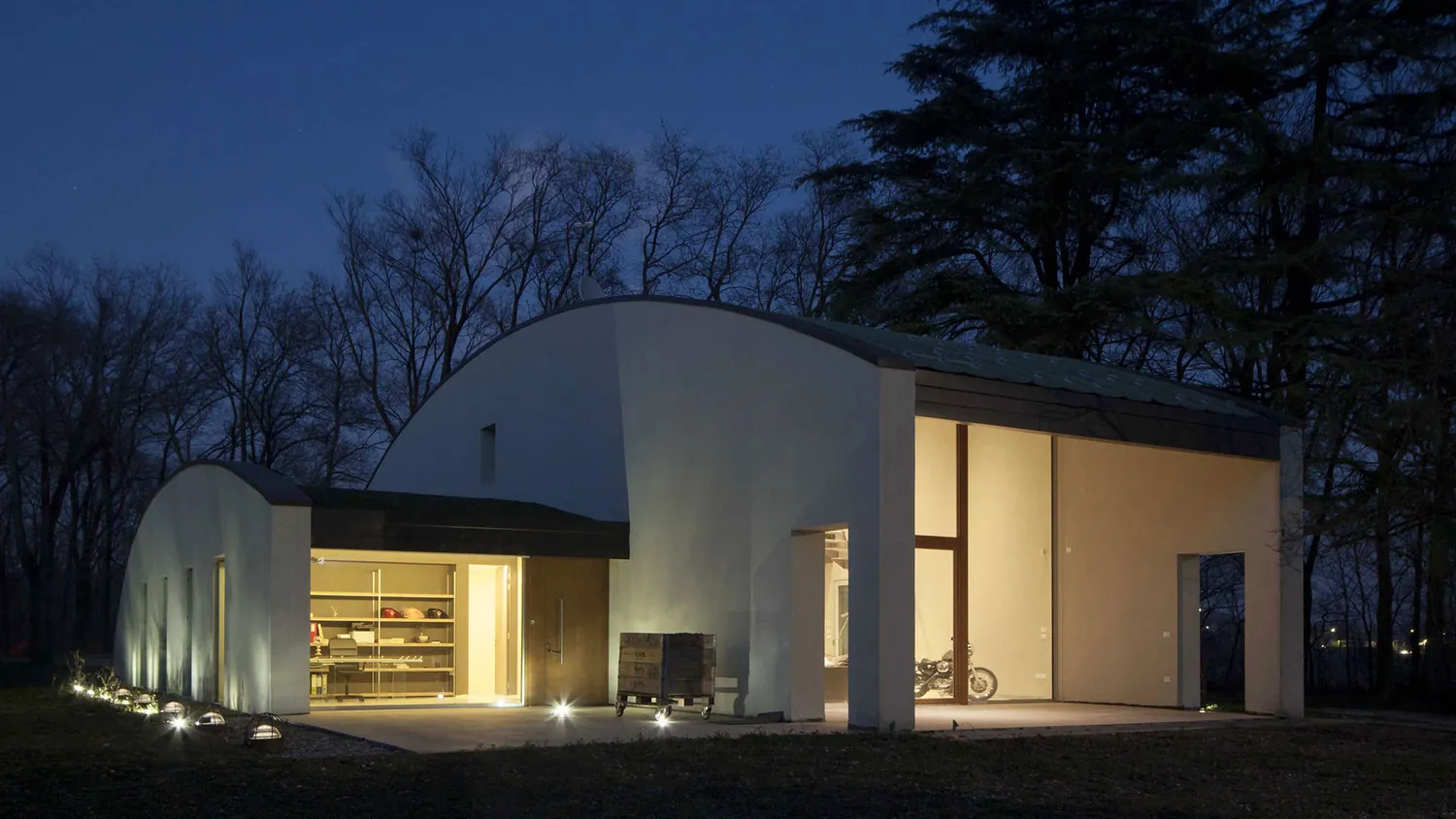 Villa in classe A+ di sera, con la copertura curva e le grandi aperture illuminate tra gli alberi