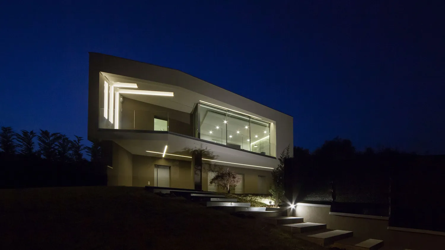 La villa illuminata di notte, con i tagli luminosi in facciata e le scale nel prato