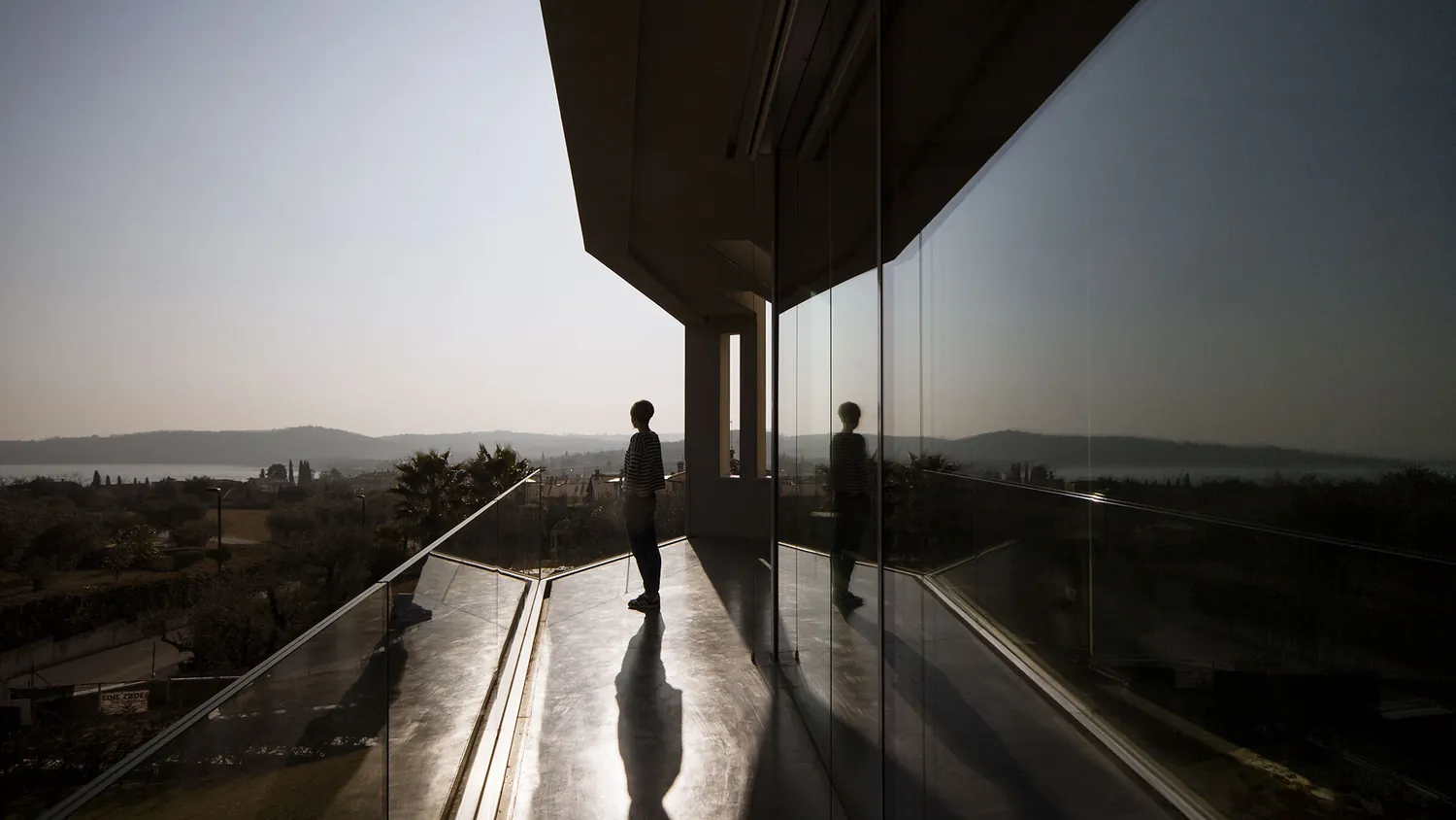 Terrazza con parapetto in vetro e una figura in controluce, con il lago all’orizzonte