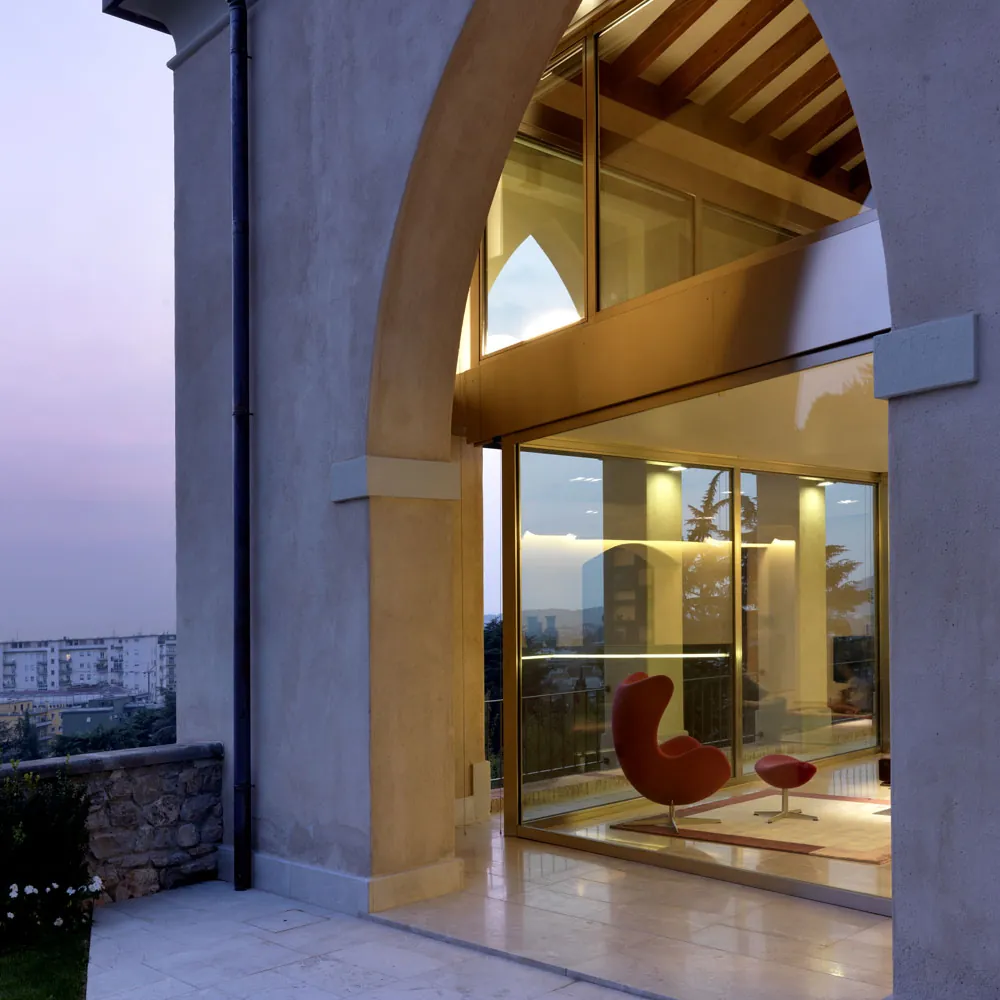 Portico ad arco di un edificio storico chiuso da una vetrata a doppia altezza, con il soggiorno illuminato e la collina sullo sfondo, al tramonto