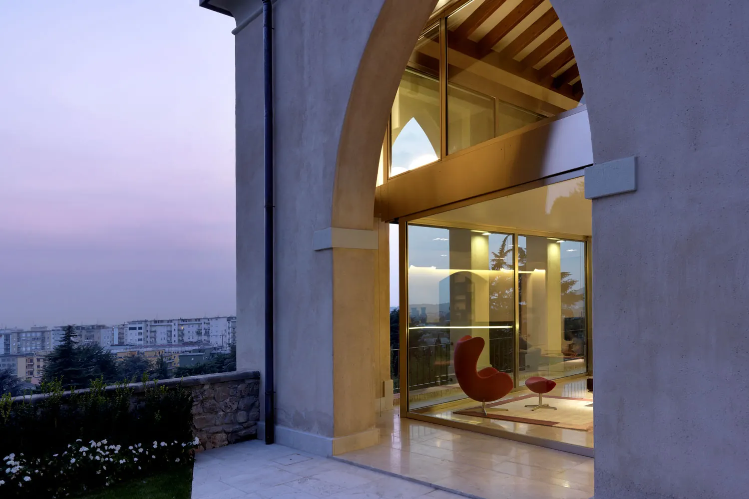 Portico ad arco chiuso da una vetrata a doppia altezza, con il soggiorno illuminato e la collina sullo sfondo, al tramonto